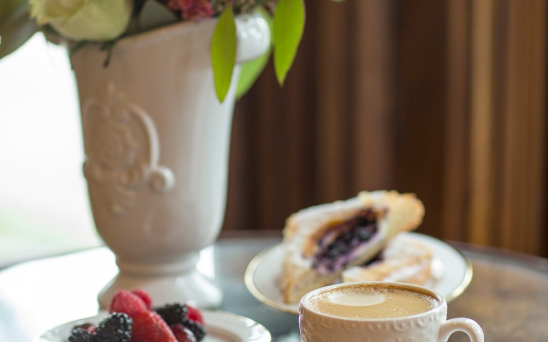 An ornamental coffee cup filled with creamed coffee on a napkin on a saucer. Beside and behind it is a plate of fresh fruit, a berry scone, and a tall vase of flowers.