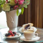 An ornamental coffee cup filled with creamed coffee on a napkin on a saucer. Beside and behind it is a plate of fresh fruit, a berry scone, and a tall vase of flowers.