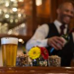 A glass of beer is on the bar with paper daisies next to it. In the background is a smiling bartender with a wine bottle in hand.