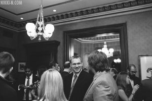 A black and white photo of people in a full room. Several ar smiling and talking. The chandeliers are bright.
