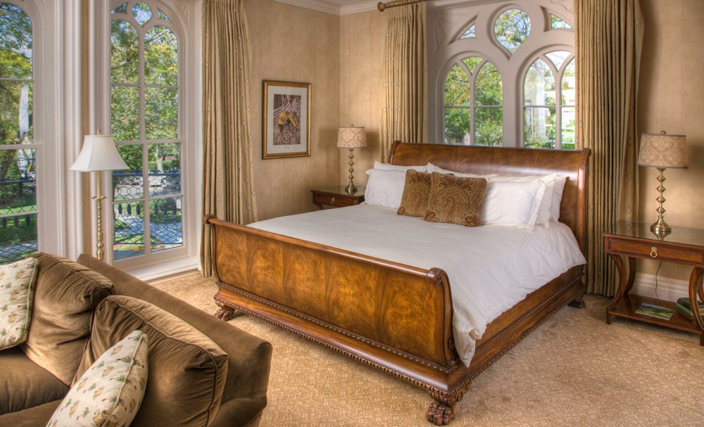 King-sized bed made up with white sheets and pillows. Two room-height windows showing greenery outside.