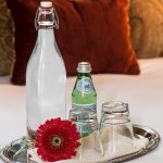 A tall bottle of still water and a small bottle of sparkling water along with two downturned glasses on a tray with a red paper daisy.