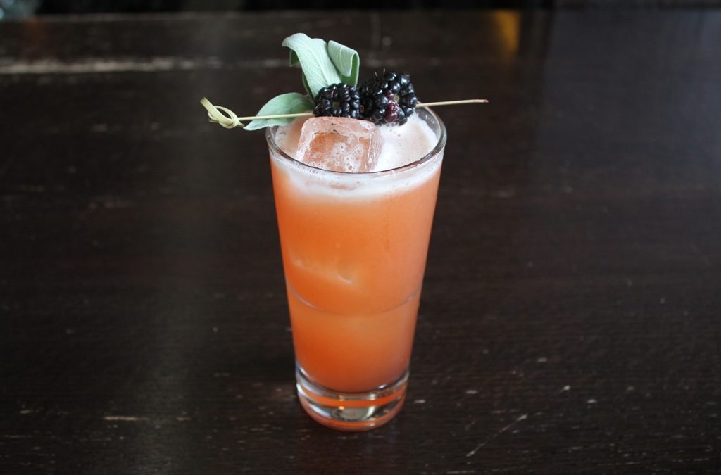 A frothy orange-pink drink in a highball glass, garnished with black raspberries and sage on a bamboo skewer.