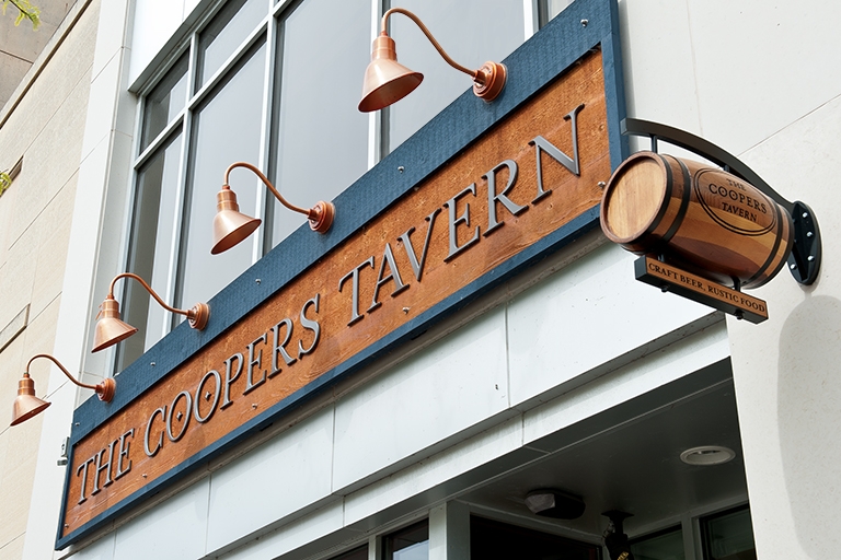An exterior sign from the street view that says "The Cooper's Tavern" with brass light fixtures above it.
