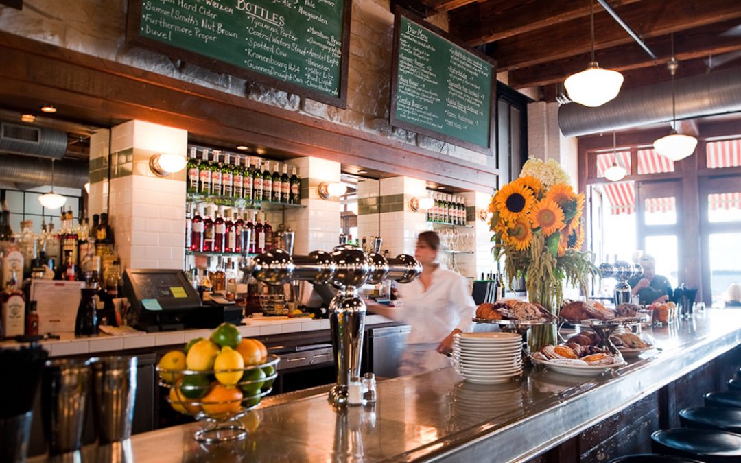 An interior view of a shiny brass bar with lots of fruit, drink accessories, baked goods, flowers, bottles of Pernod Anise and a bartender out of focus.