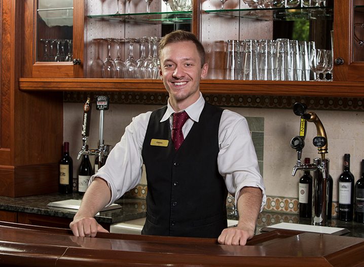 Christos, with a short cut, standing behind a bar, smiling at the camera.