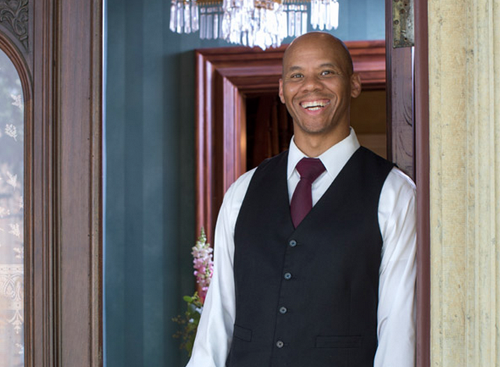 Rodney, with a shaved head, leaning against an open door frame, smiling at the camera.